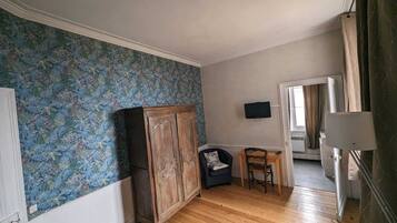 A room with wooden flooring, a wardrobe, a small table with a chair, and a television on the wall.