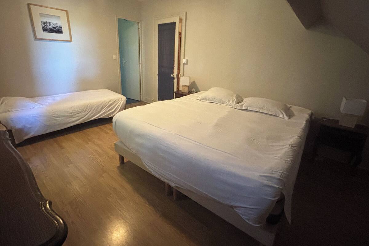 Une chambre avec deux lits simples et un lit double, un plancher en bois, un tableau encadré au mur et une porte donnant sur une autre chambre.