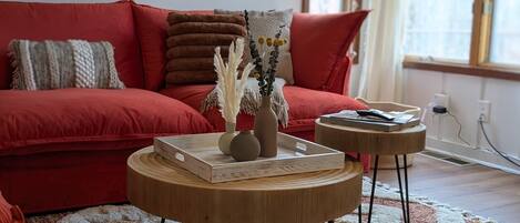 A red sofa with multiple cushions, a round wooden coffee table with a book and a vase, and a small round side table with a remote control and a book.