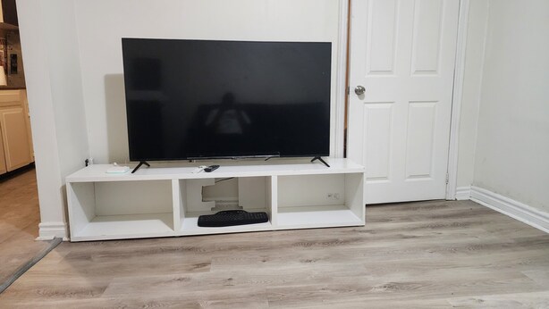 Un téléviseur à écran plat, posé sur un meuble blanc, avec un clavier et une télécommande, devant un mur blanc, dans une pièce au plancher en bois.