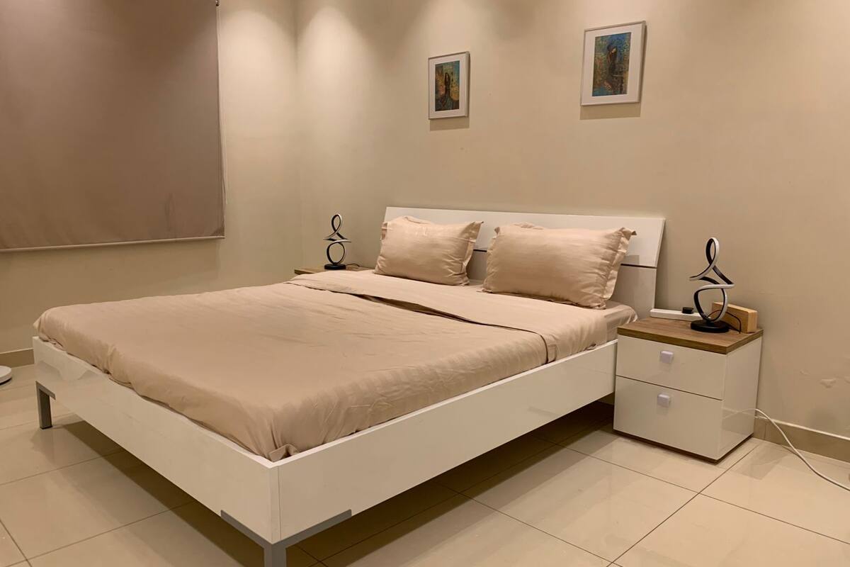 Une chambre à coucher moderne avec un lit blanc, une literie beige et deux tables de chevet dotées de supports métalliques décoratifs.