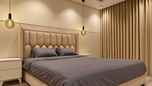 A modern bedroom with a tufted headboard, a large bed, two bedside tables, and a large window with vertical blinds. - EWA Host (Riyadh)