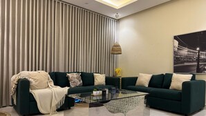 A modern living room with a dark green sofa set, a glass coffee table, and a large window with vertical blinds. - EWA Host (Riyadh)