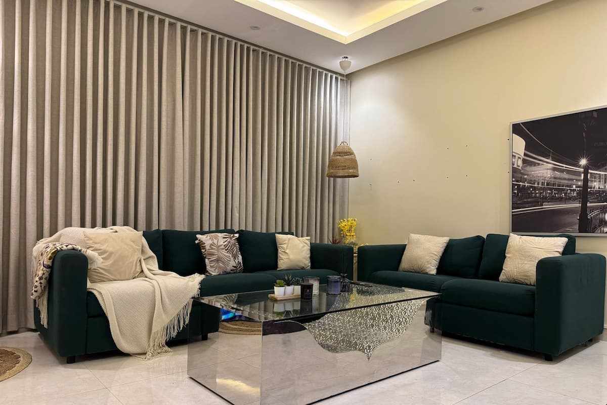 Un salon moderne avec un canapé vert foncé, une table basse en verre et une grande fenêtre dotée de stores verticaux.