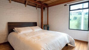 A bedroom with a wooden headboard, white brick walls, a large window with a view of greenery, and a small table. - Cari House Mountain View Near Volcano (Salinas)
