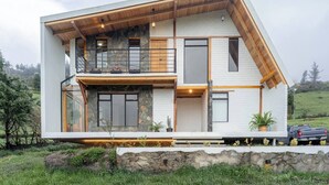 A modern two-story house with a stone facade, wooden beams, and a balcony with a glass railing. - Cari House Mountain View Near Volcano (Salinas)