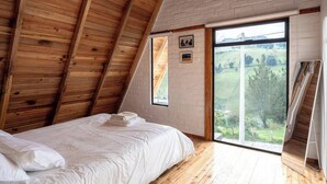 Un dormitorio con techo de madera inclinado, una cama con ropa blanca, un ventanal con vistas a un paisaje verde y un pequeño cuadro en la pared.