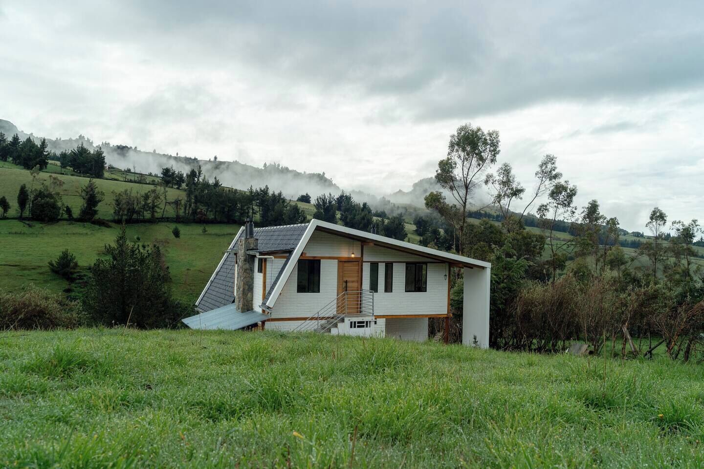 Une maison moderne avec un toit en pente, un escalier menant à l’entrée et une vue sur des collines verdoyantes en arrière-plan.