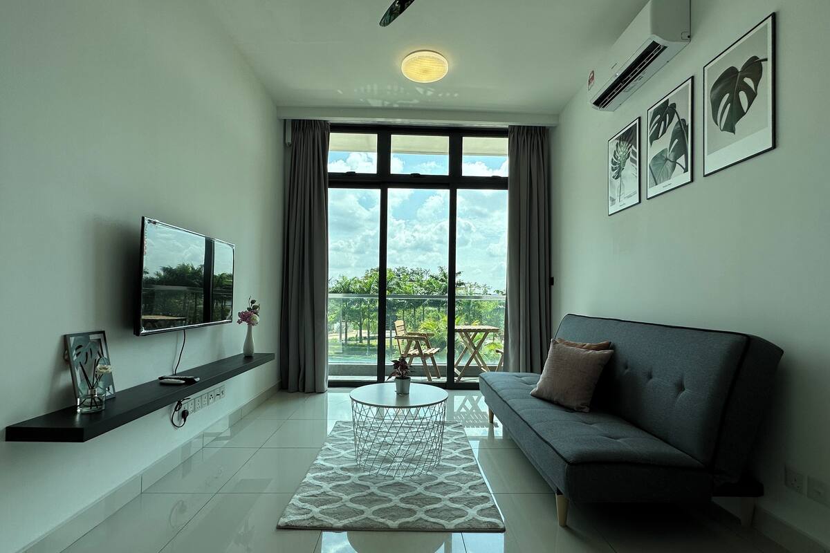 A modern living room with a flat-screen TV, a grey sofa, a coffee table, and a large window offering a view of greenery.