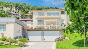 A modern two-story building with a garage, a covered outdoor seating area, and a view of a hillside with other buildings.