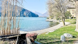 A lakeside view with a clear sky, mountains in the background, and a building with a sloped roof. - Apartment Svasso by Interhome (Lago di Mezzola)