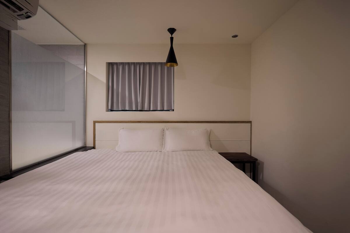 A hotel room with a large bed, a bedside table, a window with curtains, and a ceiling light.