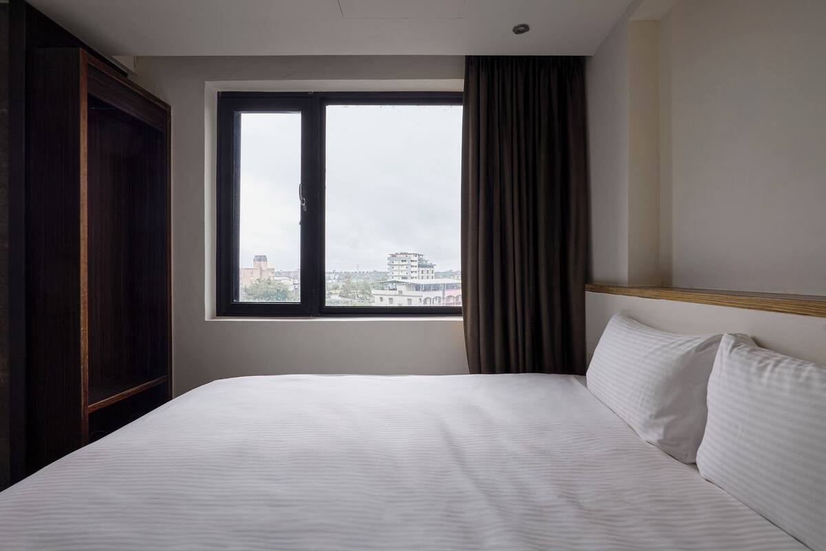 A hotel room with a large bed, a window with curtains, and a view of buildings outside.