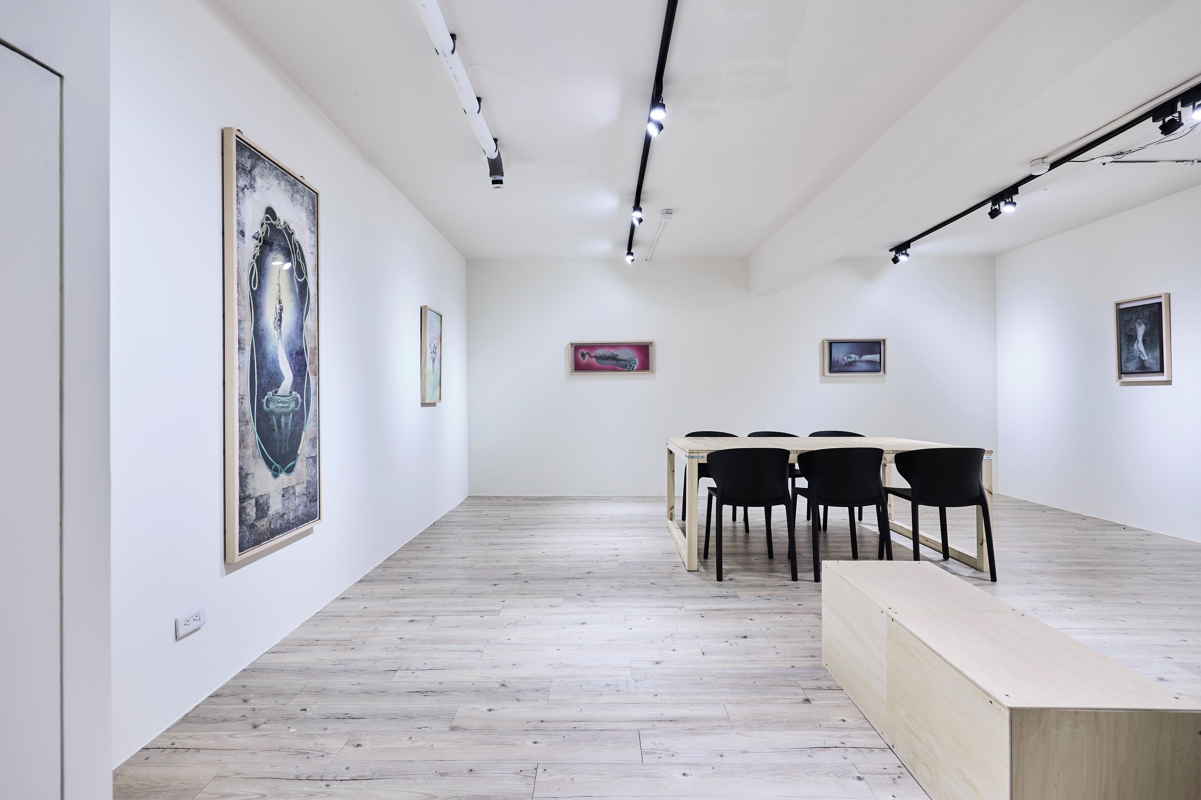 A gallery with a wooden floor, white walls, and framed artworks. There are black chairs around a table.
