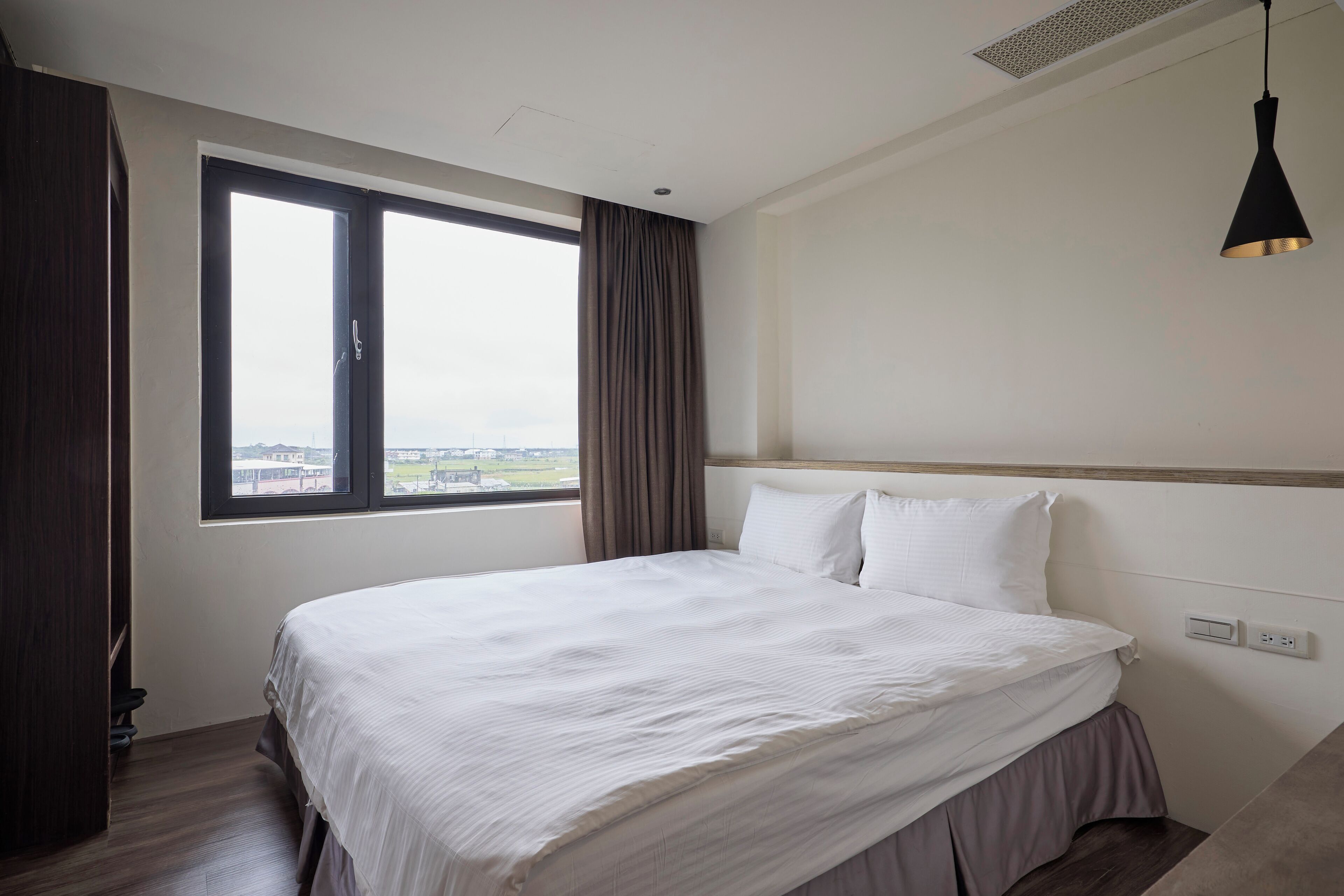 Quarto de hotel com uma cama grande, janela com cortinas, luminária no teto e vista para a cidade.