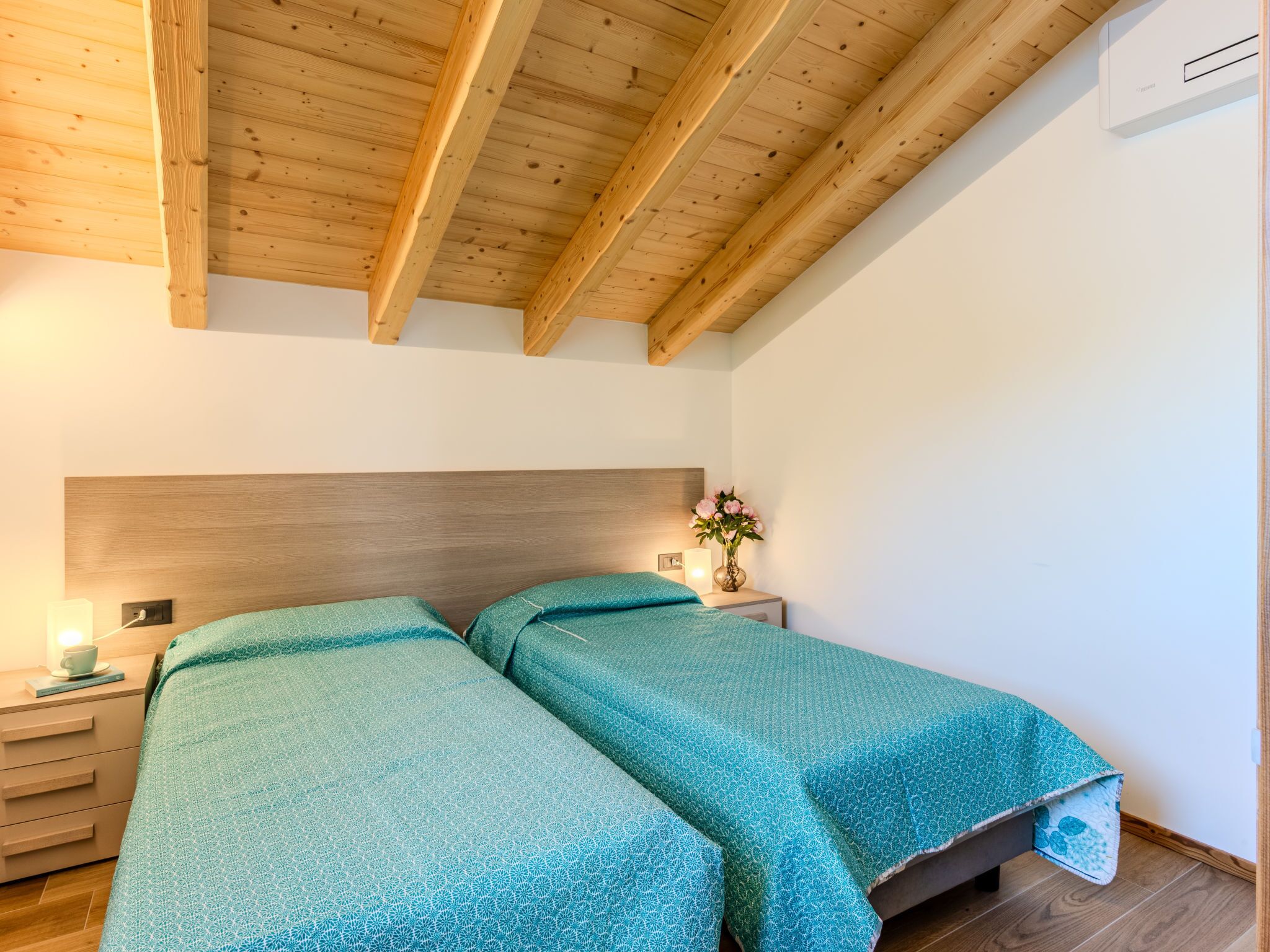 A bedroom with two beds, wooden beams on the ceiling, a bedside table with a lamp, and a vase with flowers.