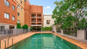Une piscine entourée d’un bâtiment en briques doté de balcons, d’un espace de repos couvert et de verdure.