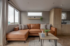 Een moderne woonkamer met een bruine leren hoekbank, een houten salontafel en een groot raam met jaloezieën.