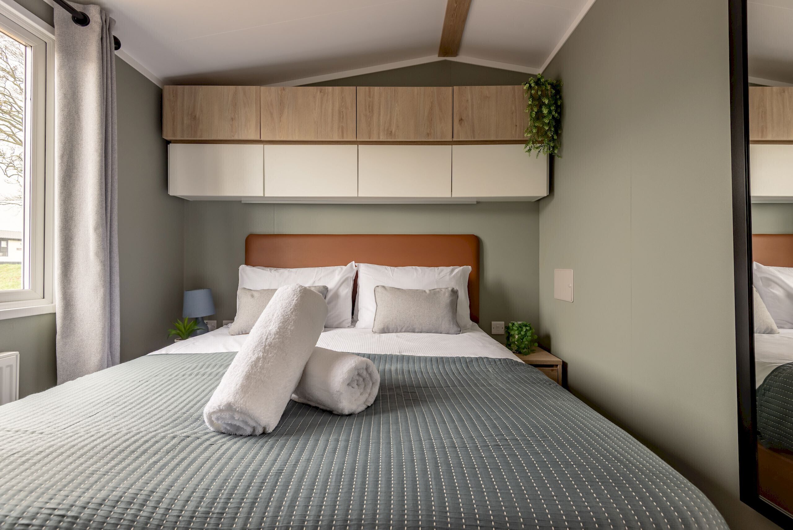 Ein ordentlich gemachtes Bett mit einer grauen Bettdecke, zwei Kissen und aufgerollten weißen Handtüchern.