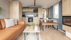 A modern living room with a sofa, coffee table, and dining area. A kitchen is visible in the background with wooden cabinets and appliances.