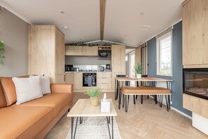 Un salon moderne avec un canapé, une table basse et un coin repas. On aperçoit une cuisine à l’arrière-plan, avec des meubles et des appareils électroménagers en bois.