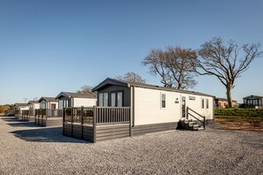 Een rij mobiele woningen met een grindpad, houten omheiningen en een grote boom op de achtergrond.