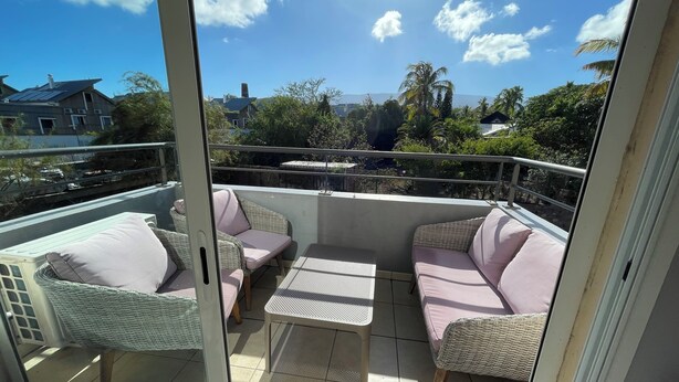 A balcony with wicker furniture and a table, overlooking a residential area with houses and trees.