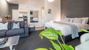 Una habitación de hotel moderna con un sofá gris, una cama con ropa blanca, una pequeña zona de comedor y una cocina al fondo.