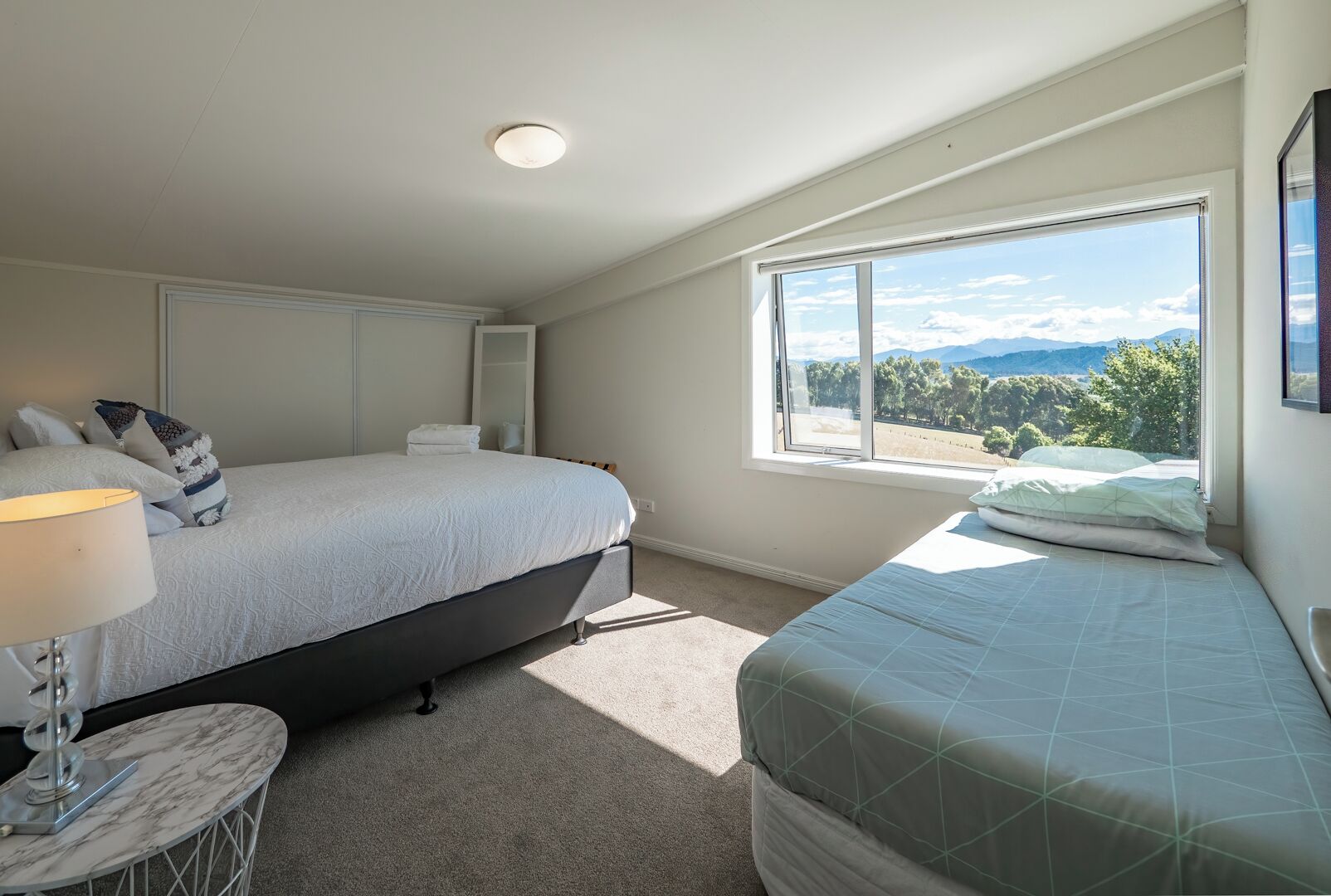 A bedroom with two beds, a bedside table with a lamp, a large window with a mountain view, and a ceiling light.