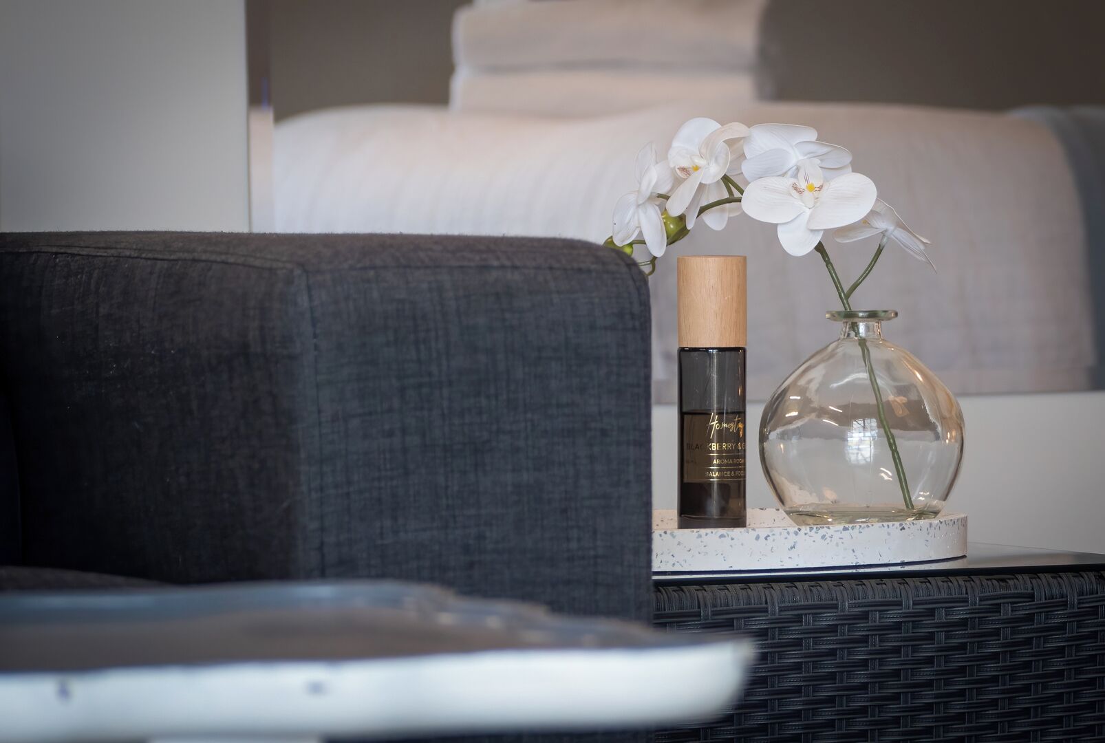 A bedside table with a white vase containing white flowers, a bottle of perfume, and a remote control.