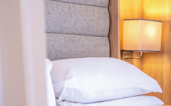 A neatly made bed with white linens and pillows, a textured grey headboard, and a wall-mounted lamp with a wooden panel.