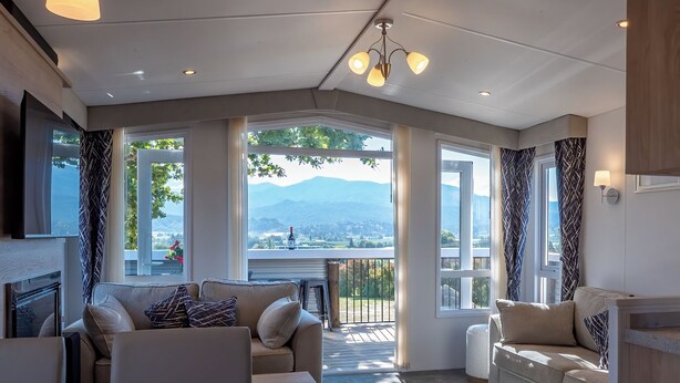 A spacious living area with a sofa set, a coffee table, and a laptop. There are large windows offering a view of mountains and a balcony.