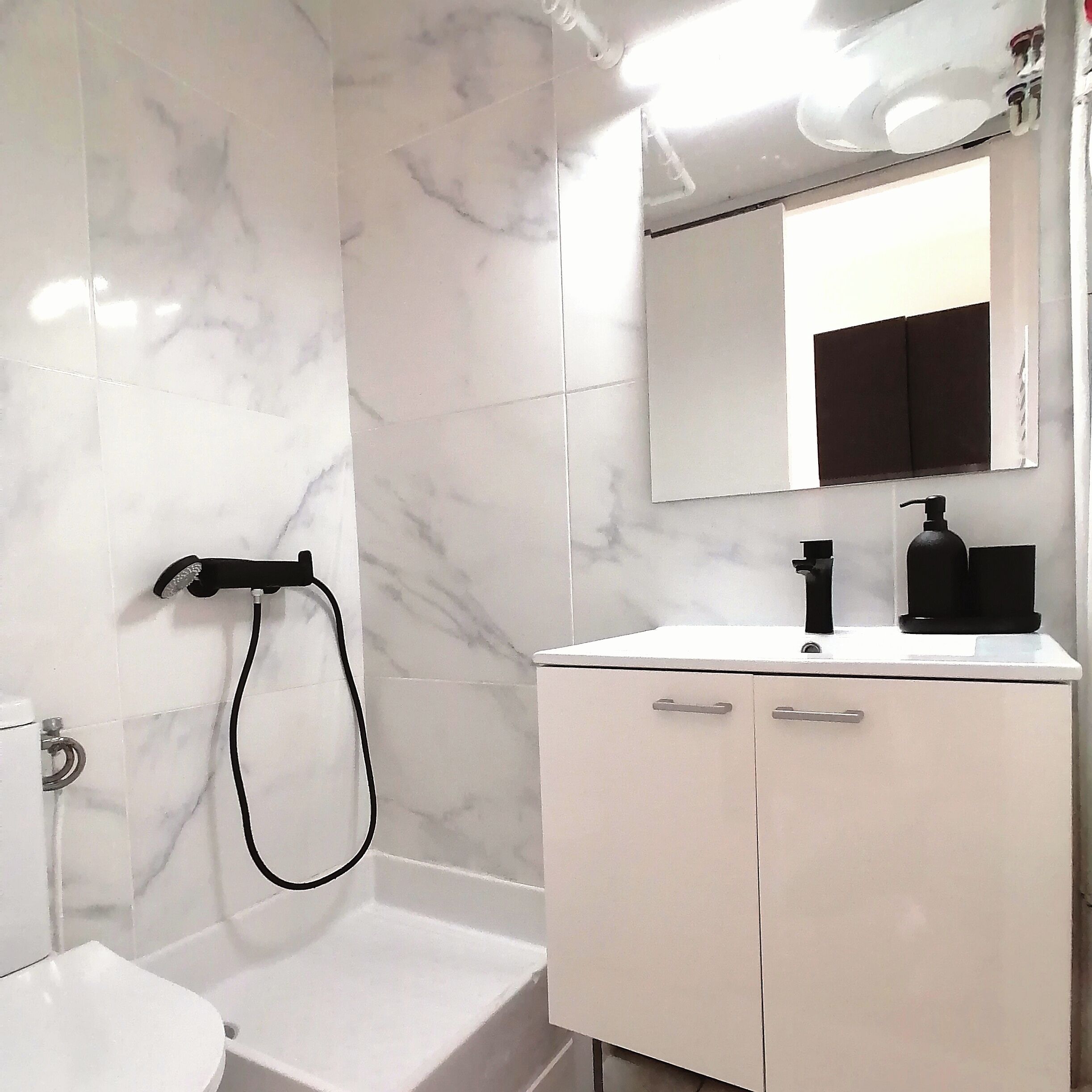 Un baño con pared de mármol blanco, un lavamanos blanco con grifo negro, un dispensador de jabón negro y un espejo sobre el lavamanos.