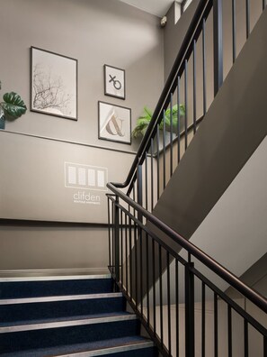 A staircase with a metal railing, a wall with framed artwork, and a logo for 'cliflen boutique apartments'. - The Town Place @ Clifden Boutique Apartments (Clifden)