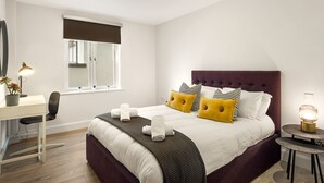 Een slaapkamer met een bed, nachtkastje, lamp, stoel en een raam met zonwering.
