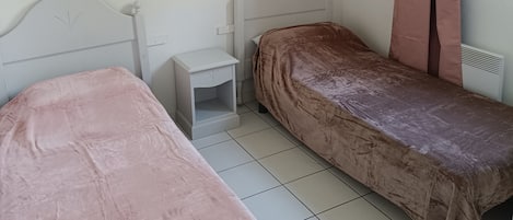 A small, simple room with two single beds, a nightstand, a hanging light, and a window with curtains.
