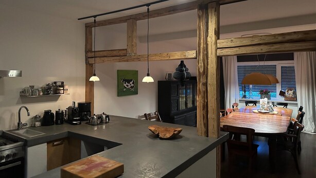 Una cocina con una isla central, vigas de madera, lámparas colgantes y vistas al comedor a través de un ventanal.