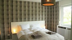 Un dormitorio con cabecera capitonada, cama con ropa blanca, dos toallas dobladas sobre la cama y una ventana con luz natural.