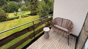 Un balcón con una silla de mimbre y una mesita pequeña, con vistas a un jardín cuidado y un edificio lejano.