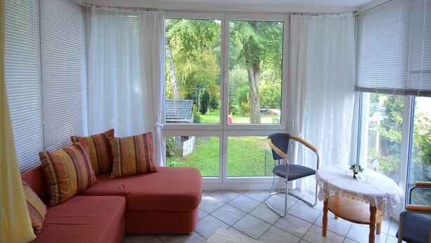 A room with a red sofa, a small round table, and a chair. There are large windows with white curtains, and a view of a garden outside. - Schönow Forest Apartment (Berlin-Schönow)