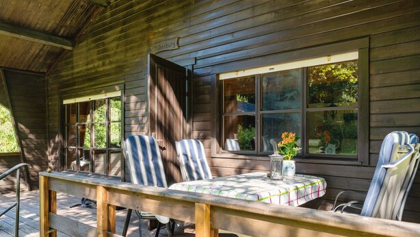 A wooden cabin with a porch, striped chairs, a table with a checkered tablecloth, and a vase with flowers.