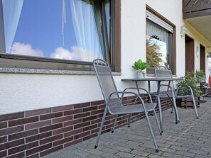 Een terras met metalen stoelen en een tafel, een bakstenen muur en een raam met gordijn.