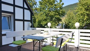 An outdoor deck with a table and benches, surrounded by a white fence, overlooking a forested hillside.