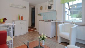 A modern kitchen with white cabinets, a microwave, and a window with a green valance. There is a dining area with a red chair and a table, and a white armchair near the window.
