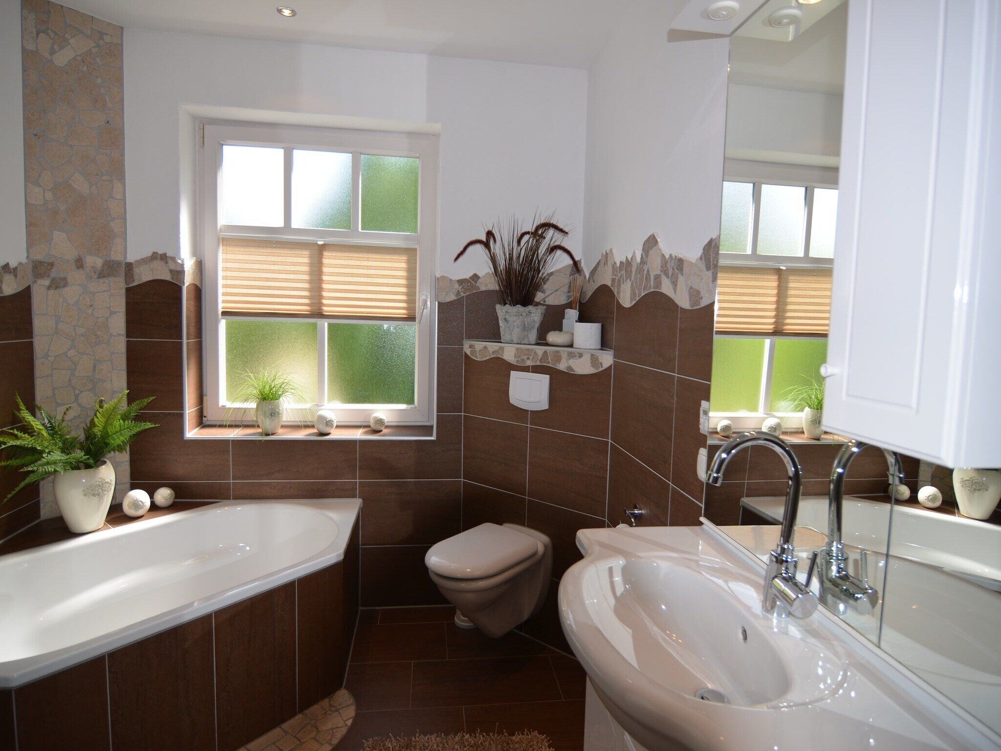 A bathroom with a large bathtub, a sink, and a toilet. There is a window with blinds, a shelf with plants, and a mirror above the sink.