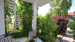 A covered patio area with a glass table and striped chairs, surrounded by lush greenery and a white staircase. - Stilvolle Wohnung in Madfeld mit eigener Terrasse (Brilon-Madfeld)