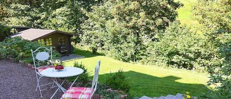 A patio with a table and chairs, a small shed, and a grassy area with trees in the background.