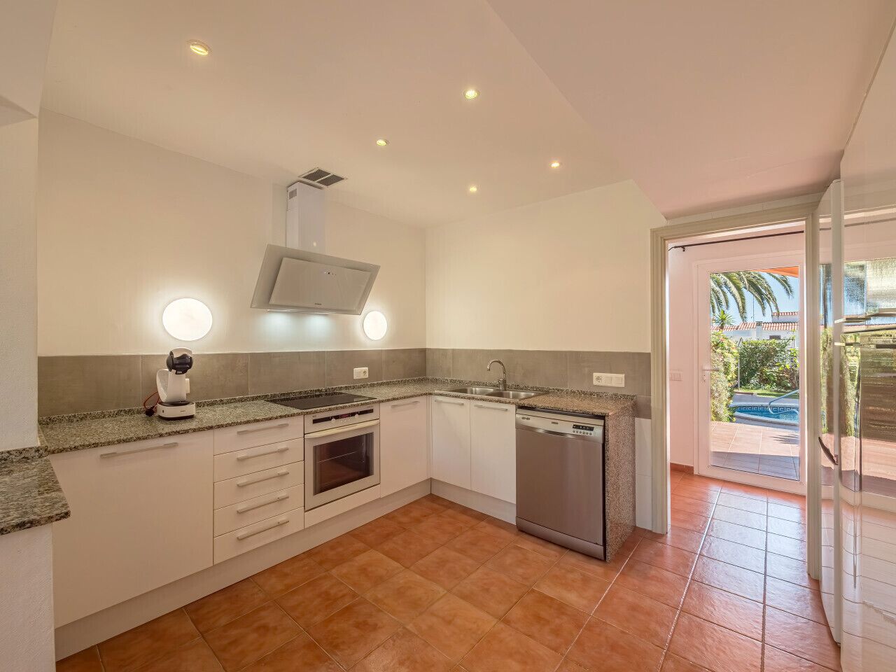 Een moderne keuken met witte kasten, granieten aanrechtbladen, een inbouwoven, een vaatwasser en een groot raam met uitzicht op een zwembad.