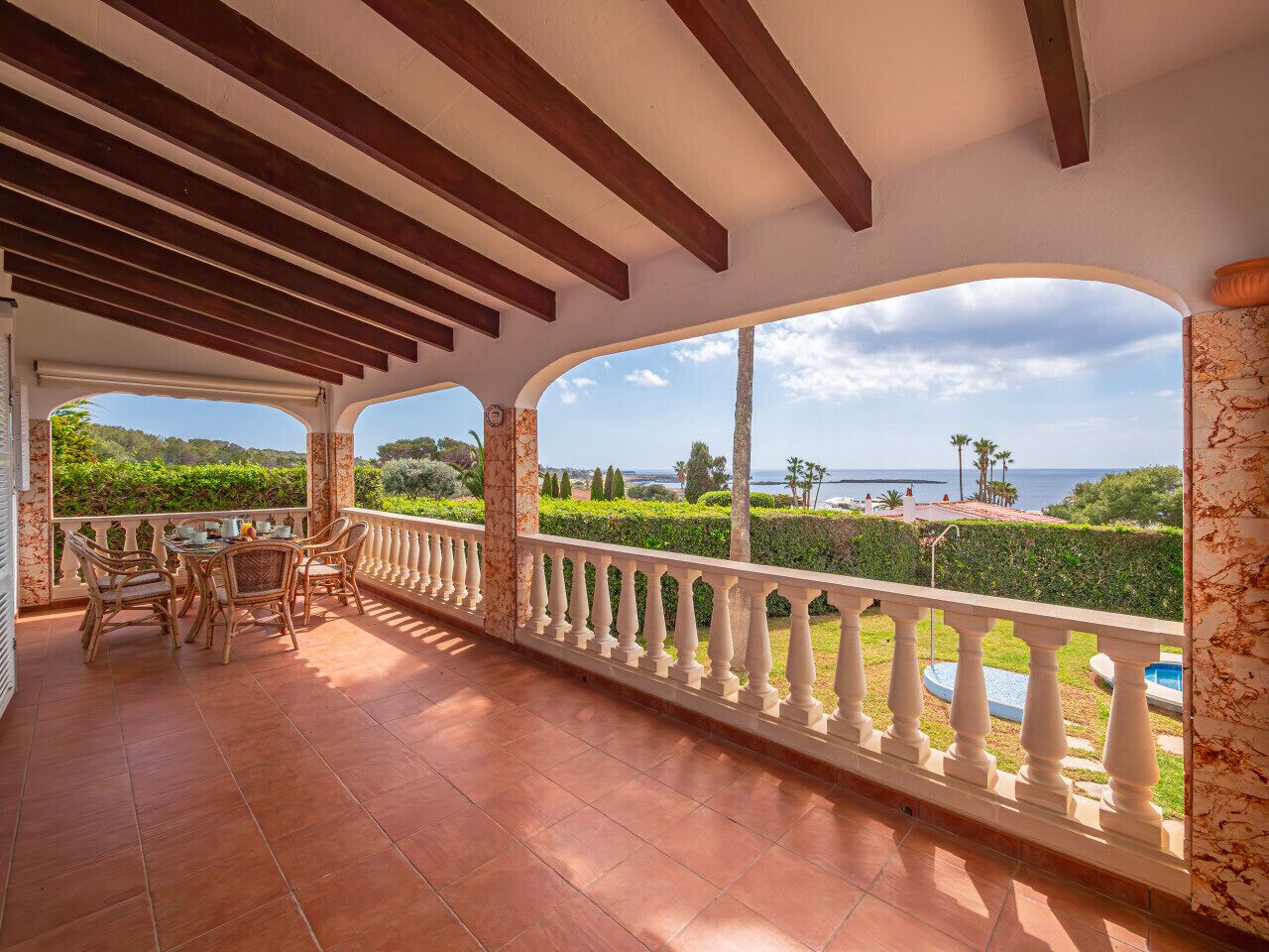 A covered outdoor patio with a dining area, white balustrades, and a scenic view of the sea and greenery.