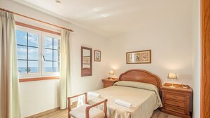 A bedroom with a wooden bed, two bedside tables with lamps, a chair, a window with curtains, and framed pictures on the wall. - Villa Sur A bit closer to heaven (Binisafua)